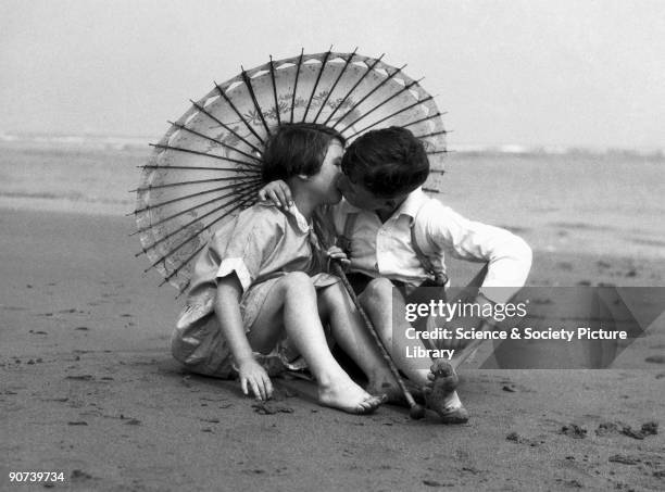 Two children on a sandy beach kissing beneath a parasol.