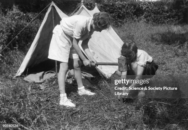 Young girl hammering a tent peg, assisted by her friend, c 1930s.