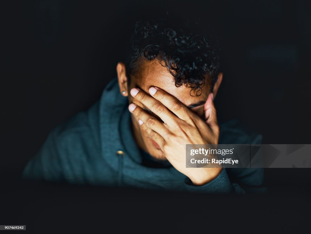 Young man sitting in the dark covers his face