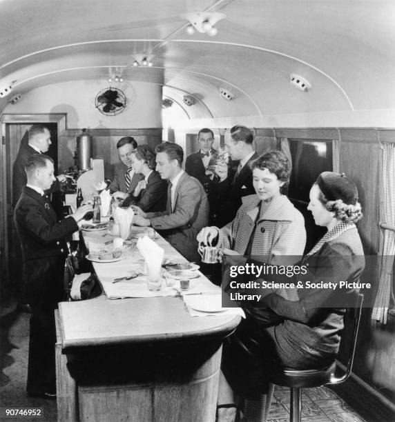 Selection of cakes, pastries and beverages are displayed on the counter. Stewards take passengers' orders for refreshments. One woman offers the...