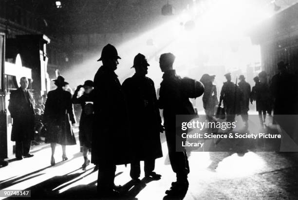 Waterloo station in wartime, London, c 1943. The roof of the station has been blacked out and the lamps shaded as hurrying figures are silhouetted...