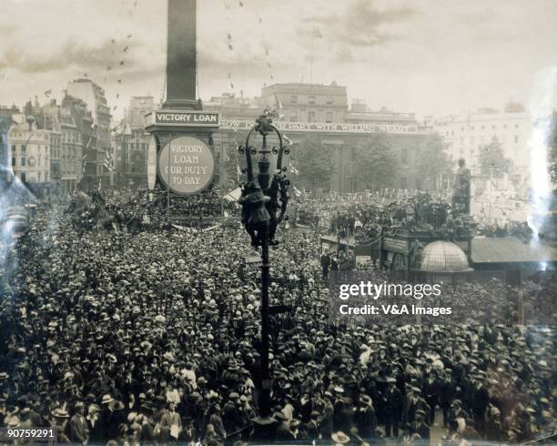 Photograph by Horace W Nicholls of crowds celebrating the end of the First World War. Hoardings round Nelson's Column read: 'Victory loan our duty...