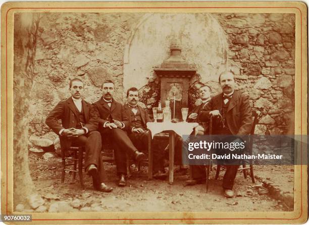 Photograph showing a group of men sitting around a table with a bottle and glasses. Behind is a fountain providing water to mix with the alcohol.