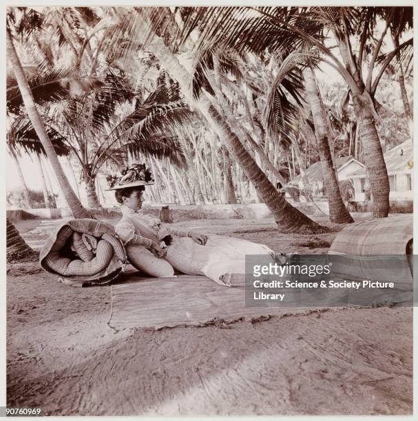 Snapshot photograph of a woman lying on the ground in the shade of a group of palm trees, taken by an unknown photographer in about 1911. Wearing...