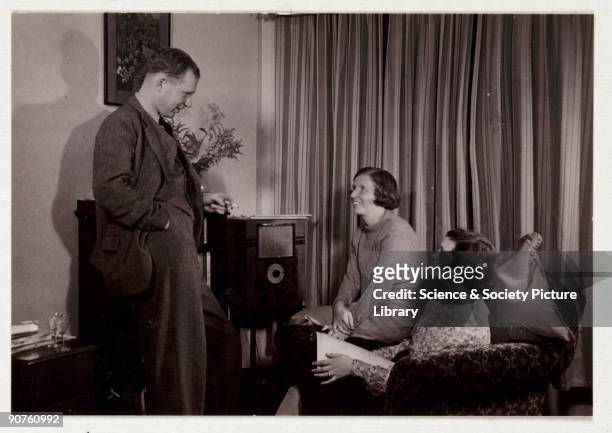 Snapshot photograph of a man and two women listening to the radio in a living room, taken by an unknown photographer in about 1935. Originally a...