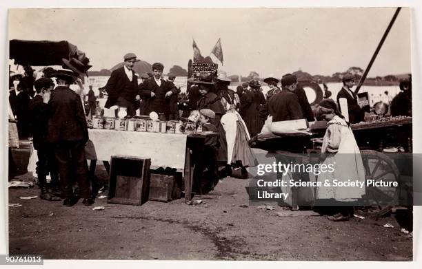 Photograph taken at a fair by Paul Martin in about 1905. Teenagers and children look over a stall displaying large tins of food. The stall on the...