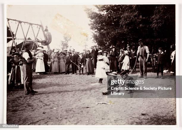 Photograph taken by Paul Martin at a fair on Hampstead Heath, London in 1898. Two men turn a skipping rope as a little girl skips. A group have...