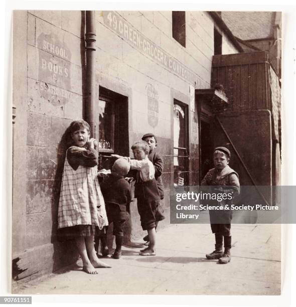 Photograph by Frank Meadow Sutcliffe of five children, some barefoot, outside a greengrocer and confectioner. The girl on the left wears a torn...