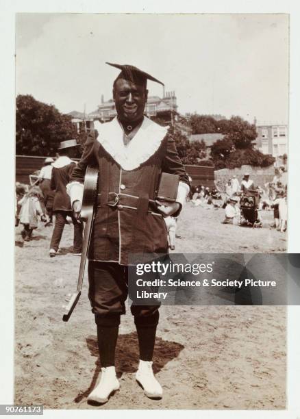 Snapshot photographic portrait of a beach minstrel with a blackened face posing for the photographer. He carries a guitar under one arm. Beach...