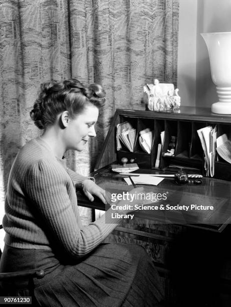 Photograph of a woman sitting writing a letter at a desk, taken by Photographic Advertising Limited in 1949. This photograph is an example of the...