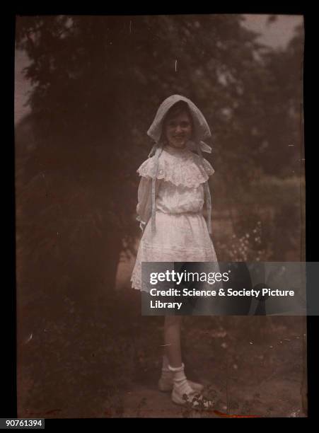 An autochrome of a smiling young girl, daughter of the photographer, wearing a bonnet and lace dress, standing in a garden, taken by Etheldreda Janet...