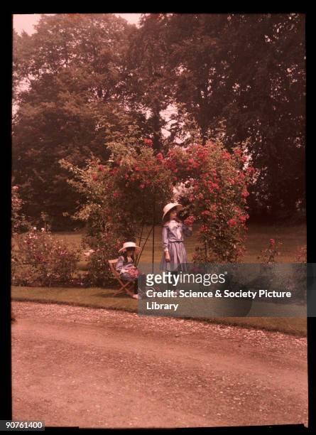 An autochrome of her daughters in a sun-filled garden, taken by Etheldreda Janet Laing. One girl picks red roses on the arched trellis. The younger...