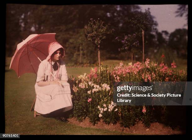 An autochrome of a girl, daughter of the photographer, with a parasol by a flower bed, taken by Etheldreda Janet Laing. In the summer of 1908 Laing...