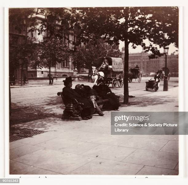 London streetscene photographed by Paul Martin [1864-1905] in about 1900. Three people rest on a bench sheltering from the sun on a tree-lined London...