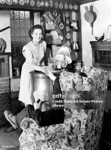 Photograph of a couple in their living room, taken by Photographic Advertising Limited in about 1950. The woman is serving the seated man a drink...