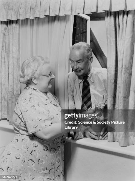 Photograph of an elderly couple talking at a window, taken by Photographic Advertising in about 1950. The image suggests contentment in retirement....