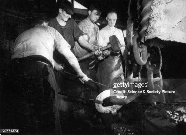 Photograph shows one man holding a white hot link of the chain with iron tongues as two other men are about to hit the link with a large mallet