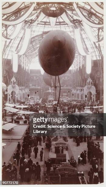 Snapshot photograph of the interior of the exhibition hall taken by an unknown photographer in 1911. The hall is dominated by an airship moored just...
