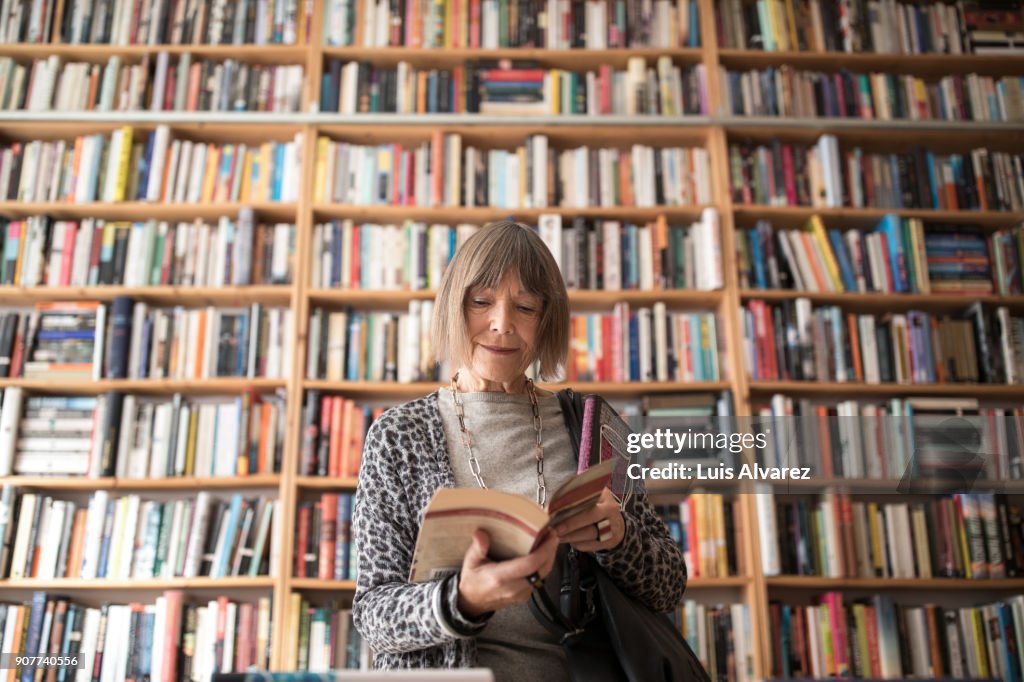 Senior woman reading book while standing against bookshelf