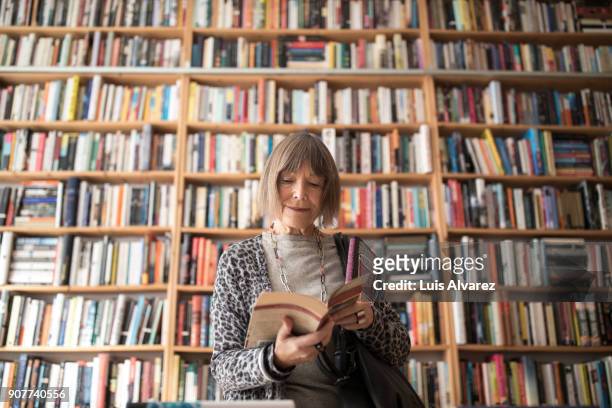 senior woman reading book while standing against bookshelf - le imagens e fotografias de stock