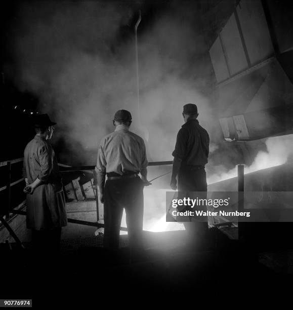 Molten steel, watched by three workers, pours from the charging end of a Carbon arc steel furnace at United Steel. The company was one of Britain's...