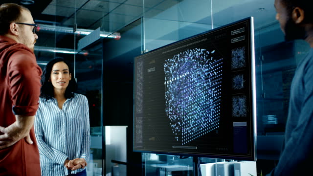 Diverse Team of Computer Engineers Discuss Neural Network Representation Shown on a Wall TV.