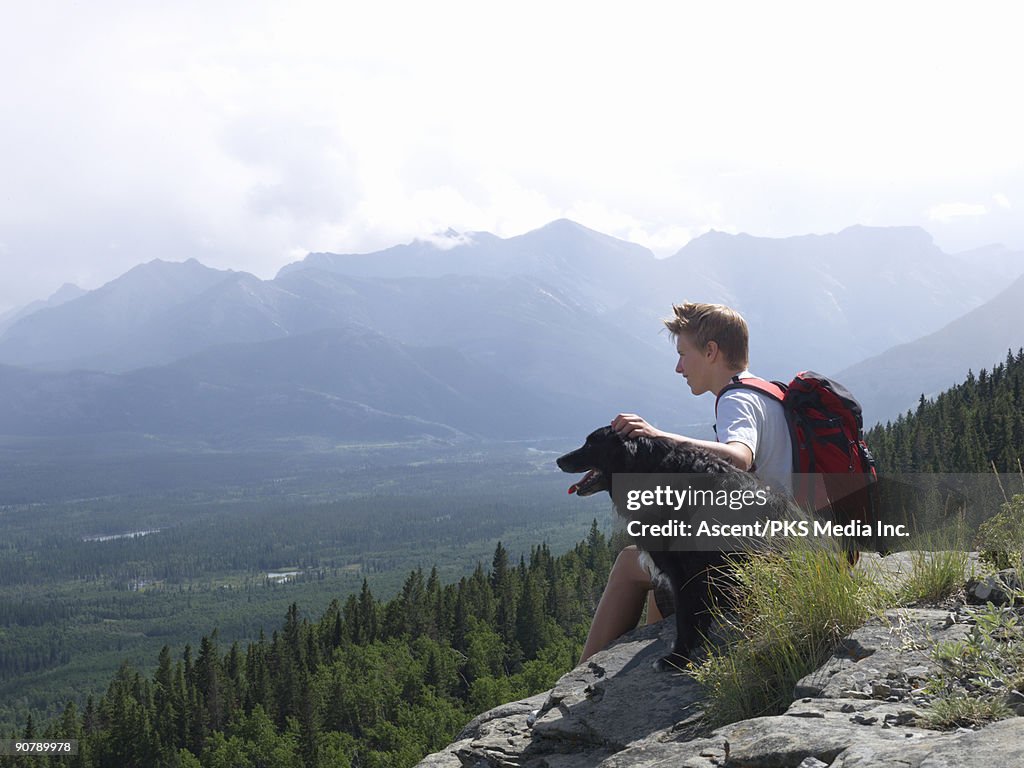 Teenage boy sits on mountain slope, pats dog