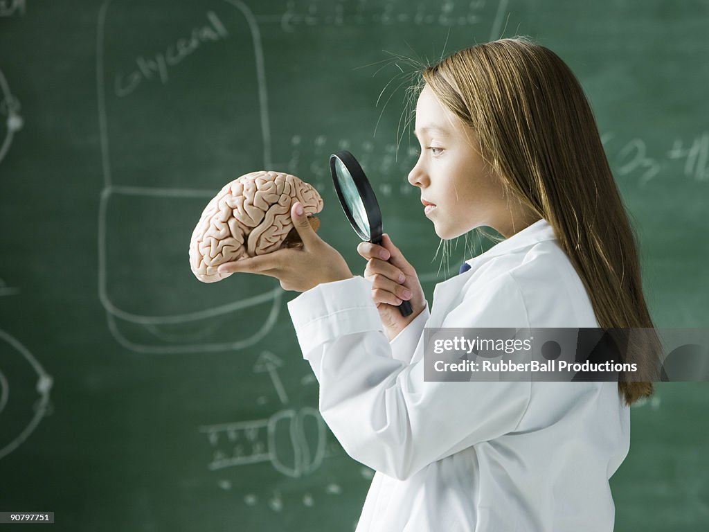 Girl in a classroom standing in front of a chalkboard looking at a brain with a magnifying glass