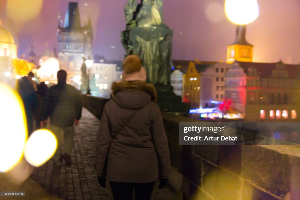 Traveler woman with winter outfit and hat visiting Charles Bridge of Prague at night with beautiful city lights and nice colors.