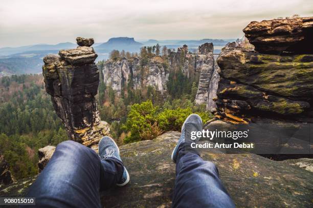 guy from personal perspective doing hiking sitting on the edge of cliff and contemplating the nice view of the beautiful saxon national park in germany with stunning views of the rock formations with vertigo and adrenaline sensations. - adrenalina fotografías e imágenes de stock