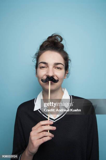 young woman in her late reens in front of a blue background holding up a fake mustache underneath her nose, portrait. - vネック ストックフォトと画像