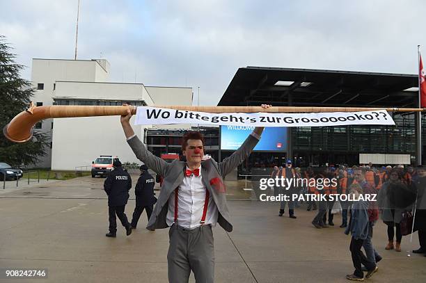 Man dressed as a clown holds up an alphorn with a banner reading "Where please is the way to Grooooooko??" as he stands in front of the venue of an...
