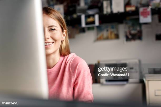 portrait of smiling young woman behind computer screen at desk at home - working at home desktop computer stock-fotos und bilder
