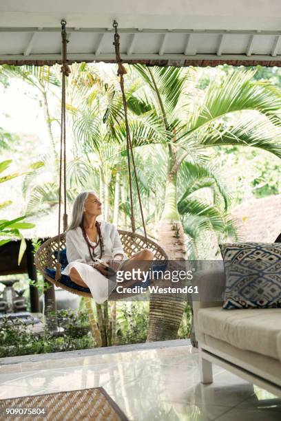 attractive senior woman relaxing in hanging chair, reading a book - hanging chair stock pictures, royalty-free photos & images