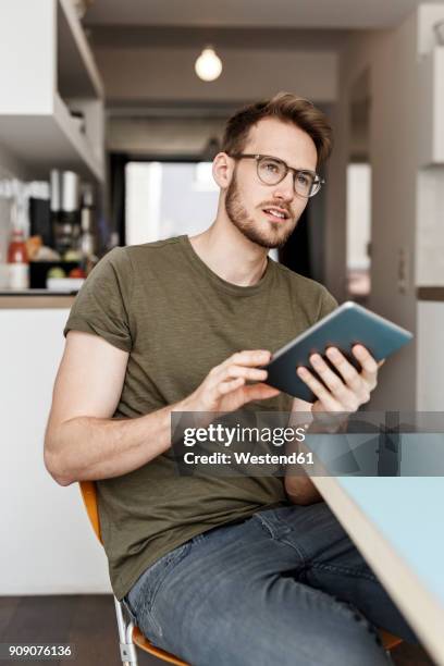 young man with tablet sitting in kitchen at home - man glasses tablet in kitchen stock pictures, royalty-free photos & images