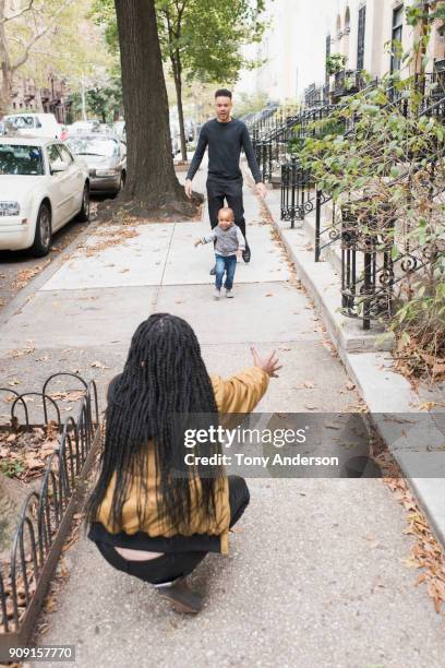 Infant daughter walking from father to mother in their city neighborhood