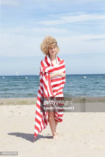 smiling young woman wrapped in beach towel standing in front of the sea - striped towel stock pictures, royalty-free photos & images