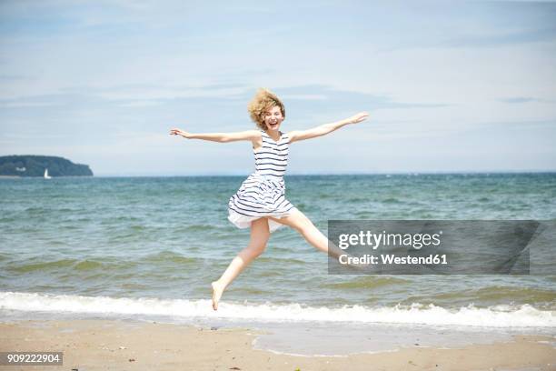 happy young woman jumping in the air in front of the sea - ringellocke stock-fotos und bilder
