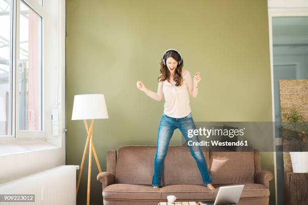 young woman with headphones standing on couch screaming and dancing - frau tanzt im wohnzimmer stock-fotos und bilder