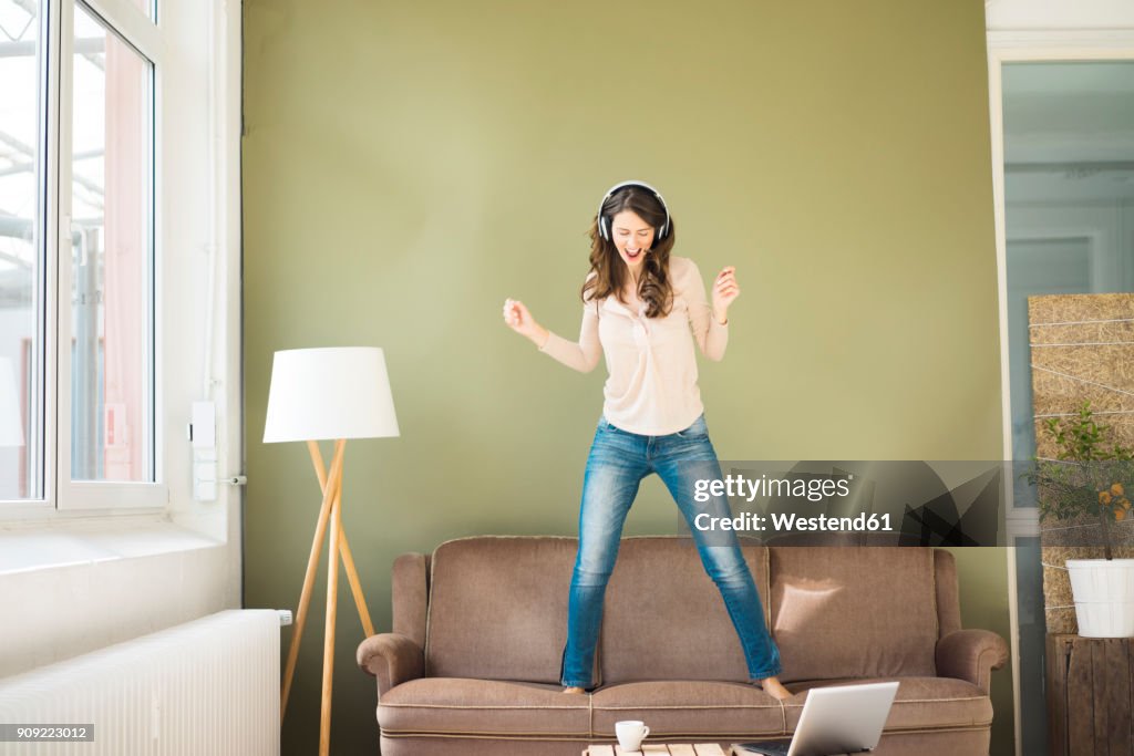 Young woman with headphones standing on couch screaming and dancing