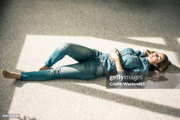 young woman lying on carpet in the living room enjoying sunlight - jeans outfit stock-fotos und bilder