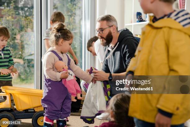 pre-school teacher helping girl putting on rainwear in kindergarten - child getting dressed stock-fotos und bilder