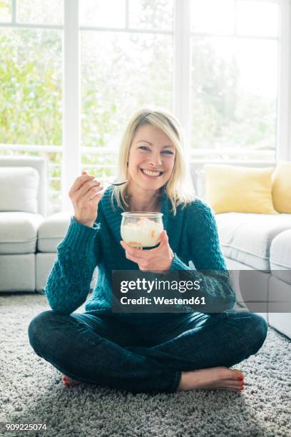 portrait of happy woman eating fruit yoghurt in living room - blond women happy eating stockfoto's en -beelden