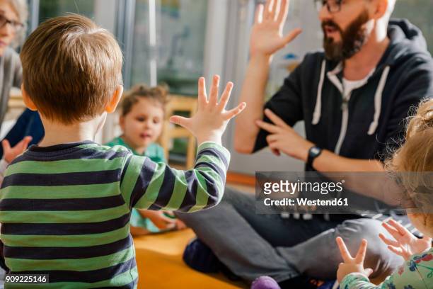 child raising hand while singing with others and teacher in kindergarten - preschool building foto e immagini stock