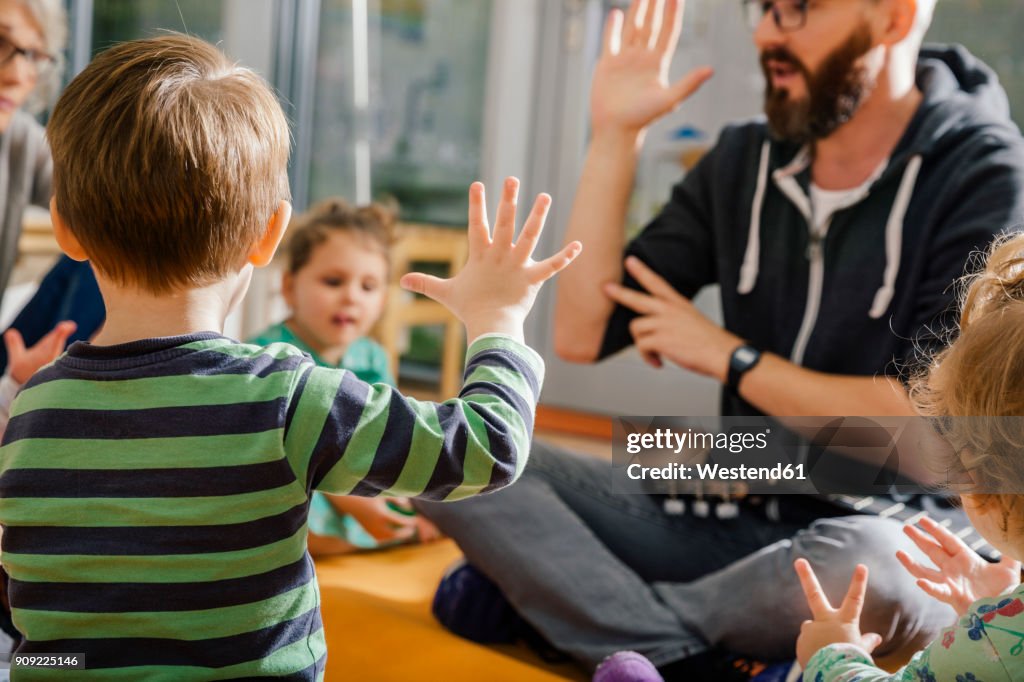 Child raising hand while singing with others and teacher in kindergarten