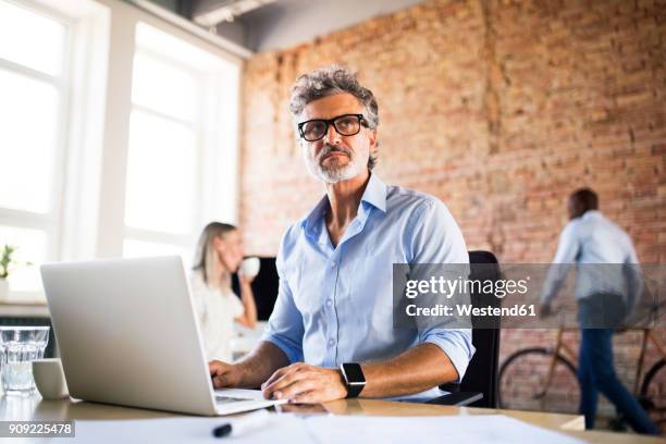 businessman using laptop in office with colleagues in background - suspicion office stock pictures, royalty-free photos & images