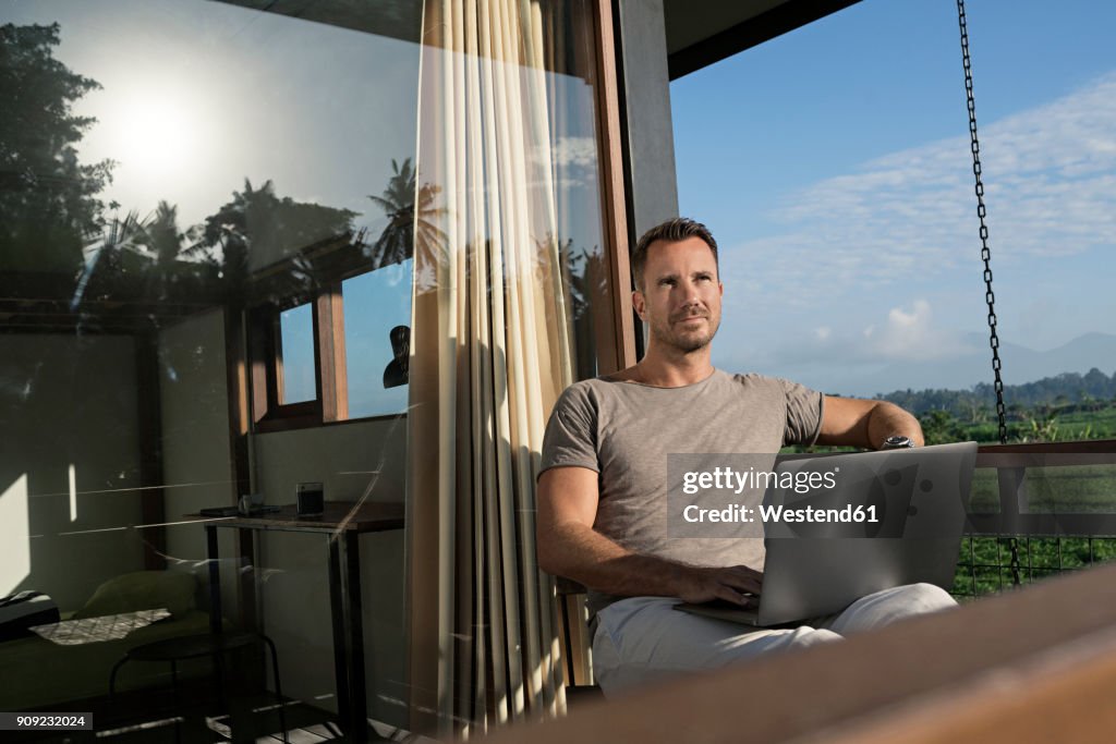Man sitting on balcony of modern house looking landscape, holding laptop