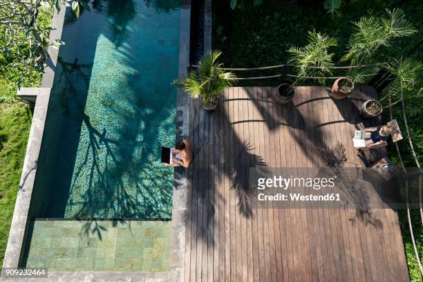 mature man sitting at the poolside, using laptop, while daughter is reading - bali luxury stock pictures, royalty-free photos & images