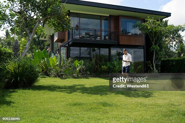mature man walking in garden in front of modern villa, using smartphone - lush grass stock pictures, royalty-free photos & images