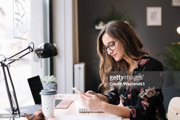 smiling woman sitting at desk in tattoo studio looking at cell phone - woman entrepreneur looking at phone stock-fotos und bilder
